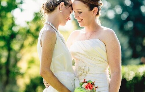 two brides look lovingly at each other, one holding a bouquet of green and red flowers